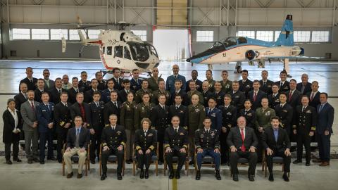 Multiple developmental test professionals pose for an official class portrait in a hangar with aircraft stationed in the background.