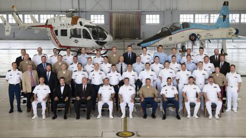Military and civilian test pilots in uniform and formal attire pose for a class photo inside a hangar with a helicopter and jet. 
