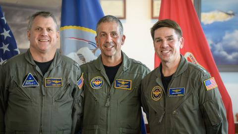 Three officers in flight suits gather for a photo in front of flags in an office setting.