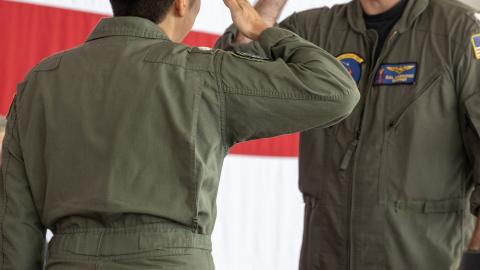 Two naval officers exchange salutes in front of a large American flag.